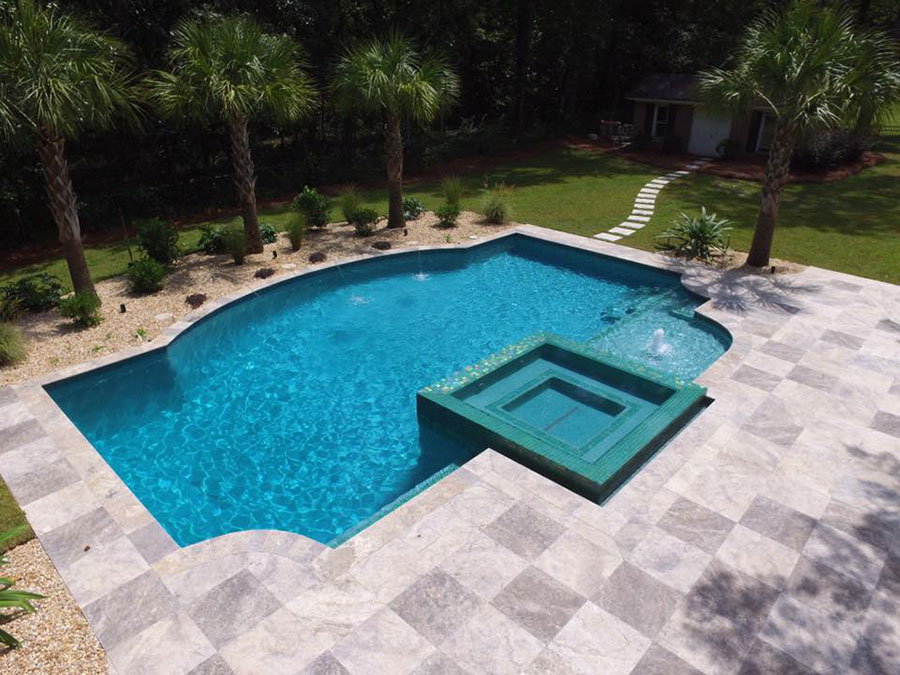 Aerial view of Mystic Pools custom freeform gunite pool in Adel with attached square spa, green glass tile accents, travertine paver decking, and tropical palm tree landscaping
