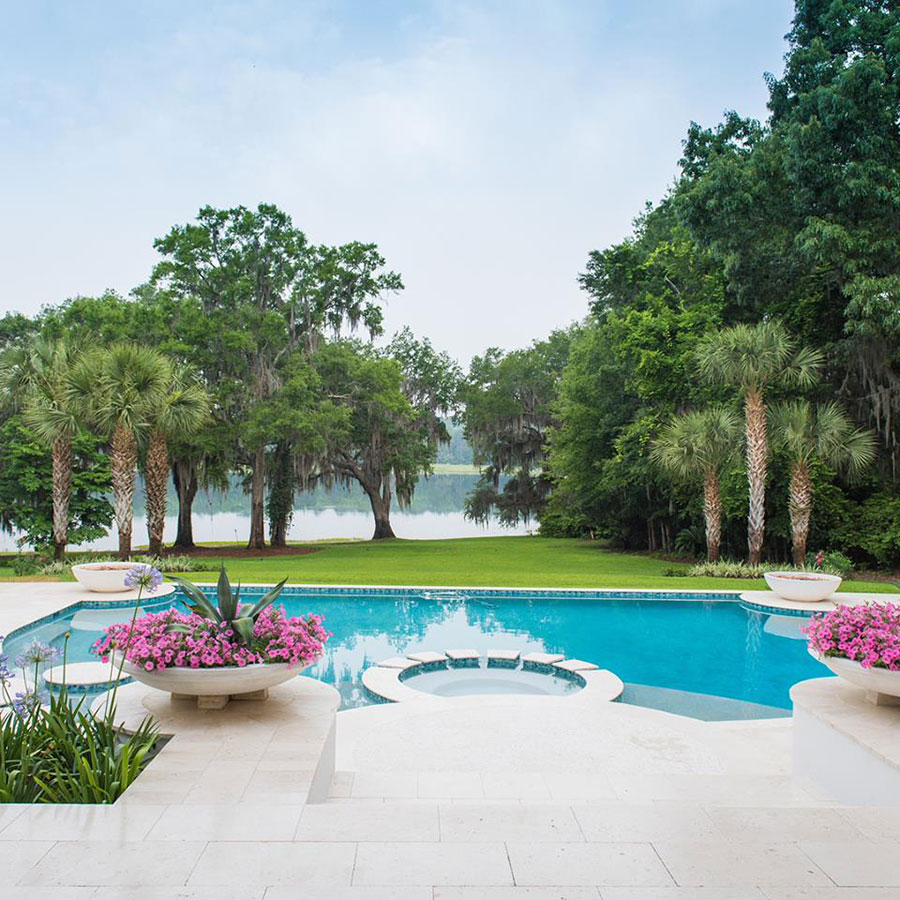 Mystic Pools custom freeform gunite pool in Adel with attached circular spa, travertine paver decking, large decorative floral planters, and a scenic lakefront view framed by Spanish moss-draped oaks and palm trees