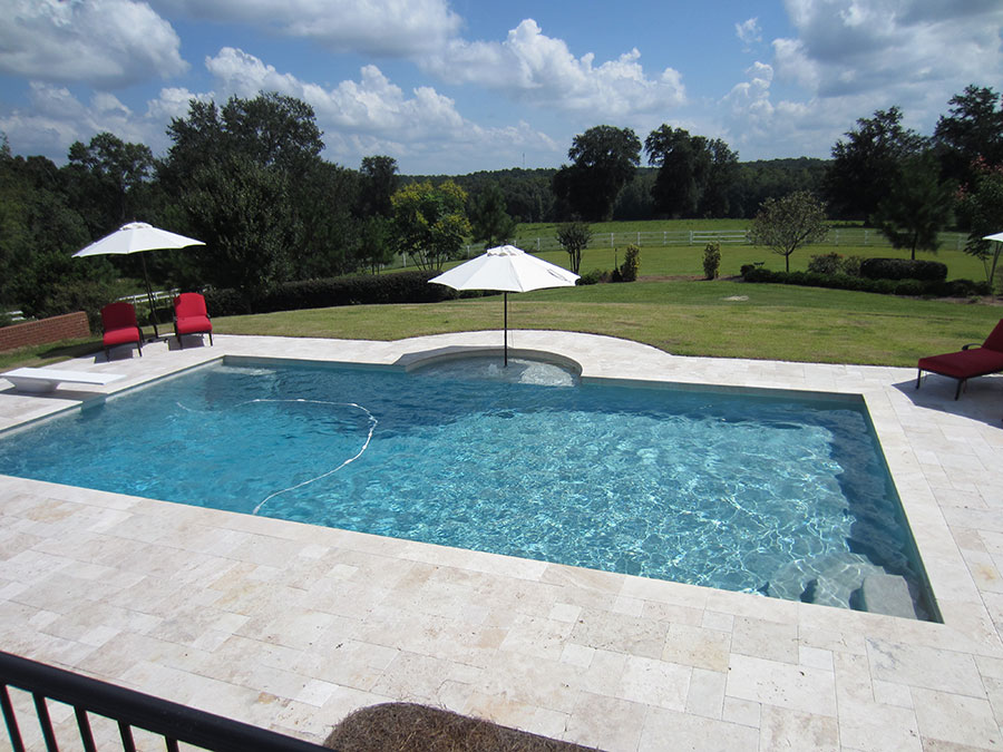 Mystic Pools custom pool design in Adel featuring a rectangular gunite pool with travertine paver decking, red lounge chairs, white market umbrellas, and a scenic view of a fenced pasture and rolling hills