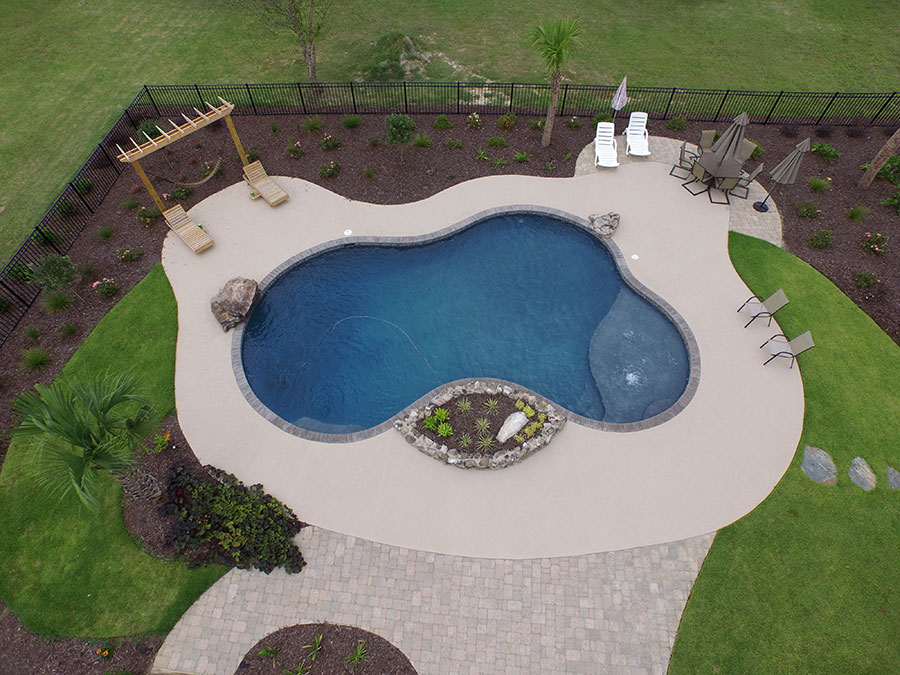 Aerial view of Mystic Pools pool renovation in Adel featuring a freeform gunite pool with submerged tanning ledge, decorative rock planter island, hammock pergola, lounge chairs, umbrella seating area, and a fenced backyard