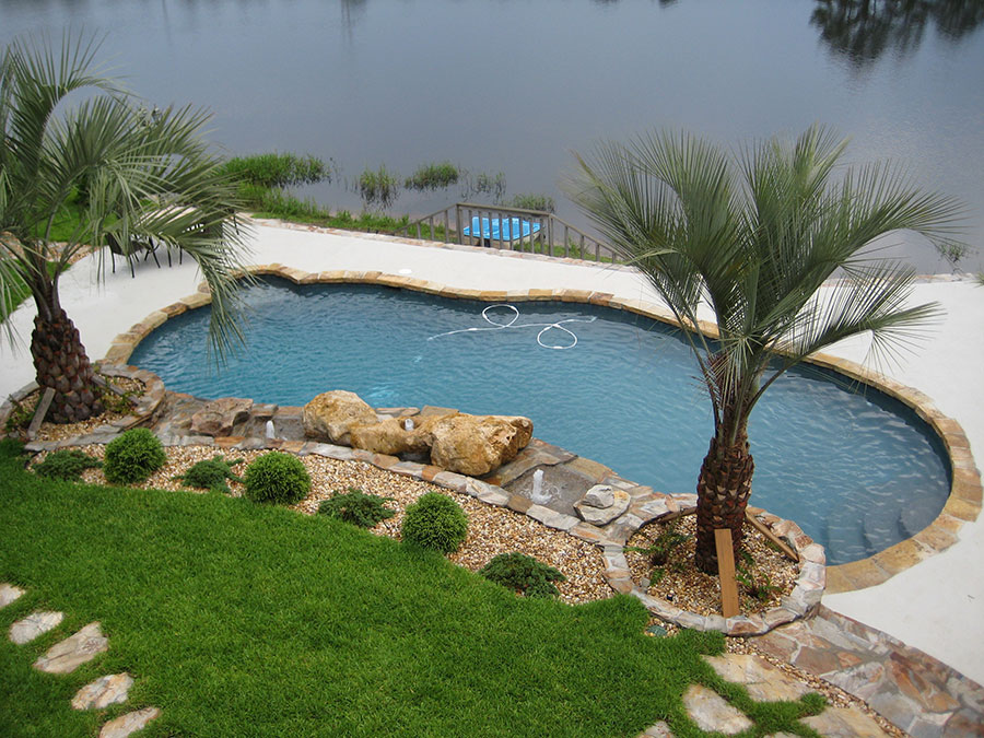 Aerial view of Mystic Pools remodeled freeform gunite pool in Adel with natural stone coping, boulder water feature, fountain jets, palm trees, gravel landscaping, and a scenic lakefront backdrop