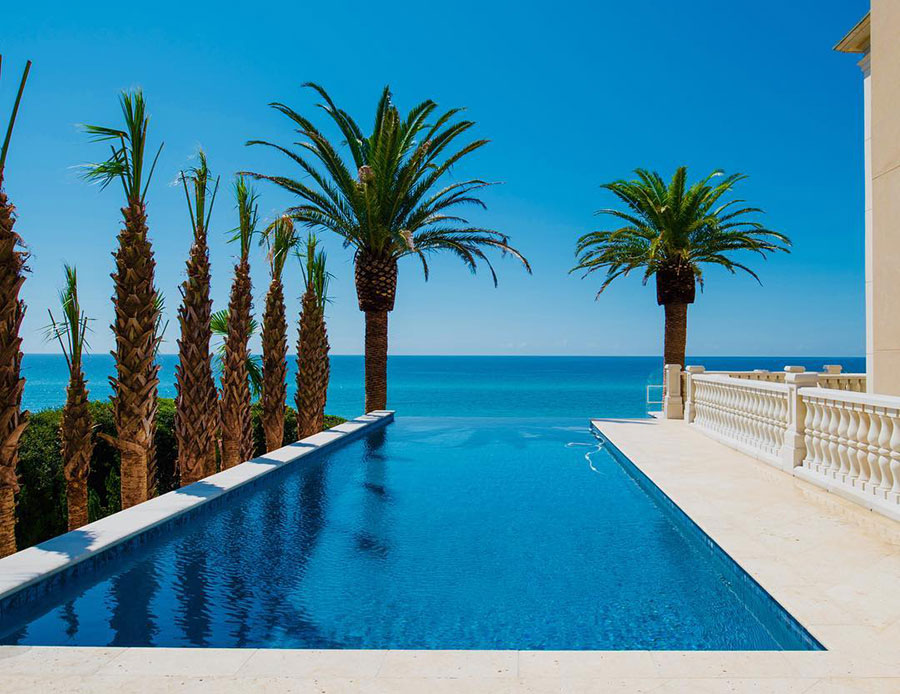 Mystic Pools custom infinity edge gunite pool in Bainbridge with dark tile finish, travertine paver decking, decorative balustrade, and sweeping ocean view framed by tall palm trees under a clear blue sky
