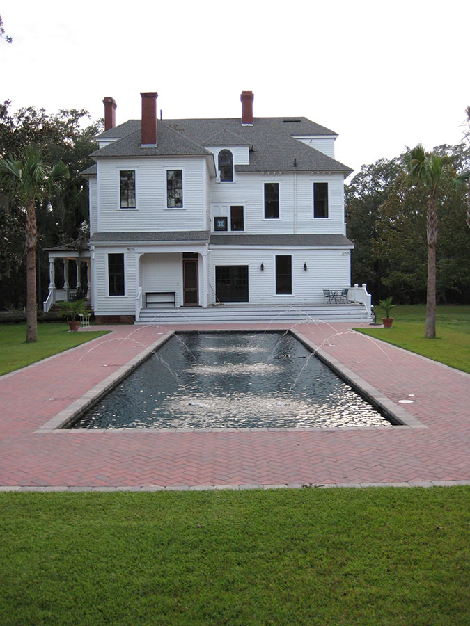 Mystic Pools custom pool design in Bainbridge featuring a long rectangular reflecting pool with multiple arc fountain jets, herringbone brick paver patio, and a historic two-story white Victorian home with palm trees