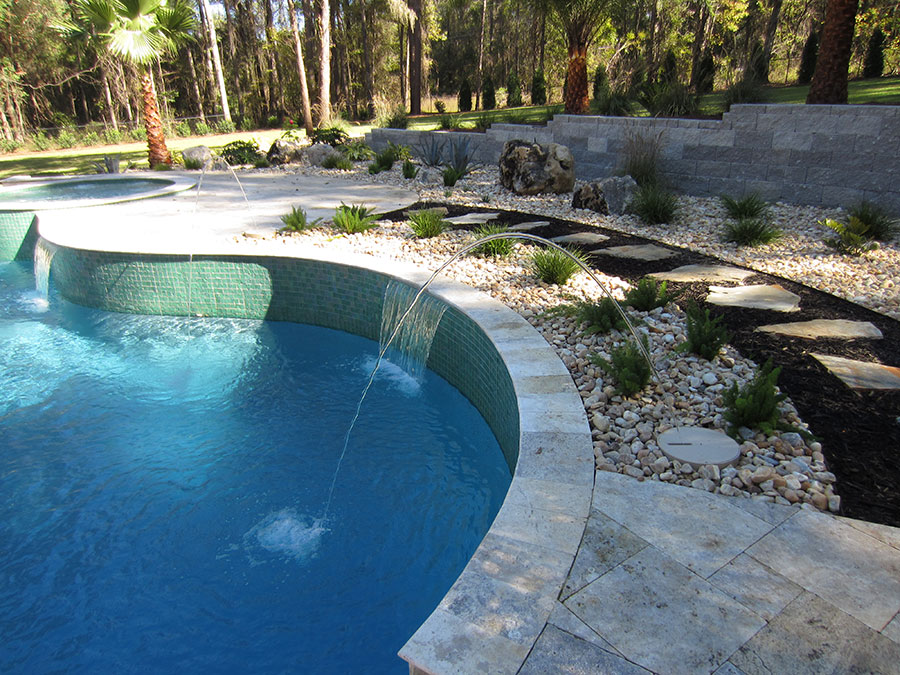 Mystic Pools custom gunite pool construction in Bainbridge featuring a curved freeform pool with green tile spillway wall, arc fountain jets, travertine paver decking, and natural stone and gravel landscaping with boulder accents