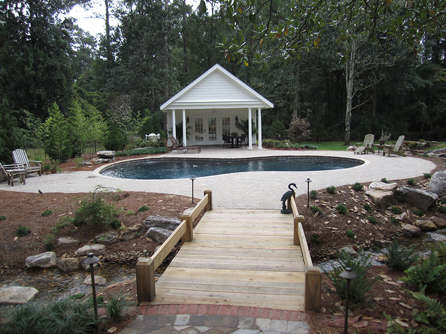 Mystic Pools pool renovation in Bainbridge featuring a freeform gunite pool with paver decking, a white pool house with covered porch, Adirondack chairs, and a wooden footbridge over a natural stream with boulder landscaping