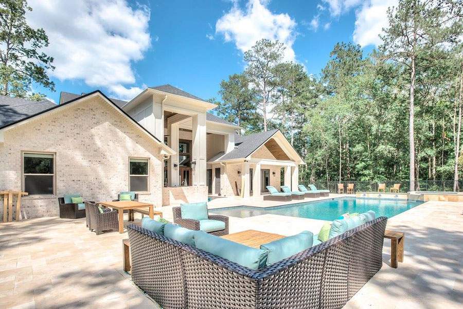 Mystic Pools remodeled rectangular gunite pool in Bainbridge with travertine paver decking, turquoise wicker sectional seating, pool house pavilion, lounge chairs, and a modern white brick estate home surrounded by tall pines
