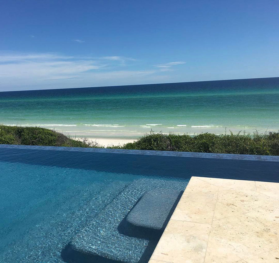 Mystic Pools custom infinity edge gunite pool in Cairo with submerged tanning ledge, dark tile finish, travertine coping, and panoramic Gulf Coast beach and ocean view