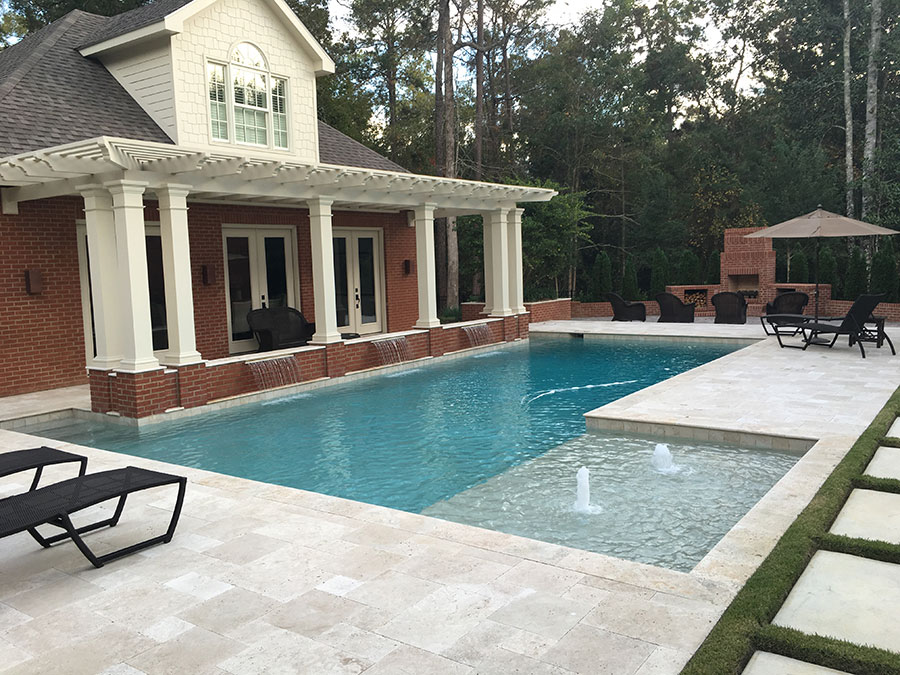 Mystic Pools custom pool design in Cairo featuring an L-shaped pool with raised wall scupper spillways, bubbler fountain jets, travertine paver decking, brick outdoor fireplace, lounge chairs, and a pergola-covered brick home