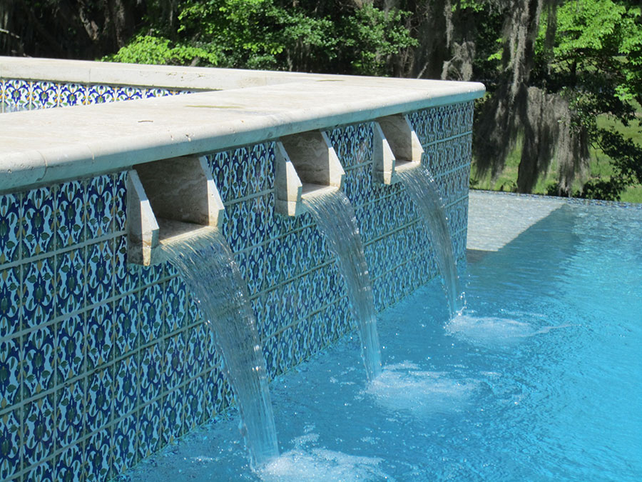 Mystic Pools custom gunite pool in Cairo featuring raised spa wall with five stone scupper spillways, ornate blue patterned mosaic tile, and travertine coping detail
