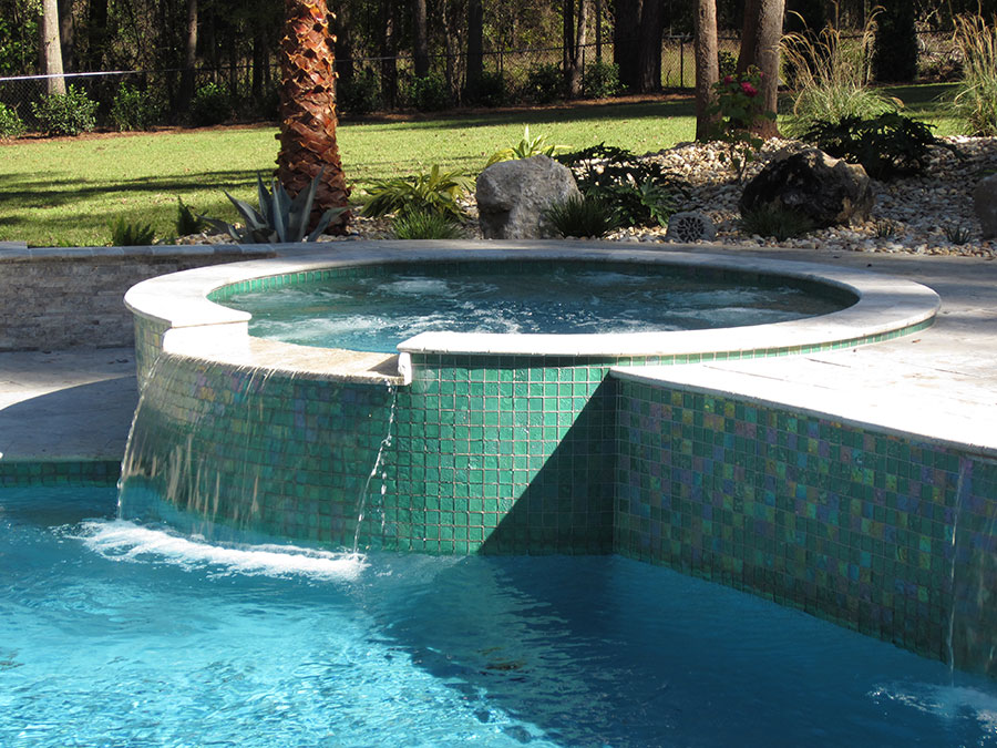 Mystic Pools custom gunite pool construction in Cairo featuring a raised circular spa with iridescent green glass tile, spillover waterfall into the pool, and natural rock and palm tree landscaping