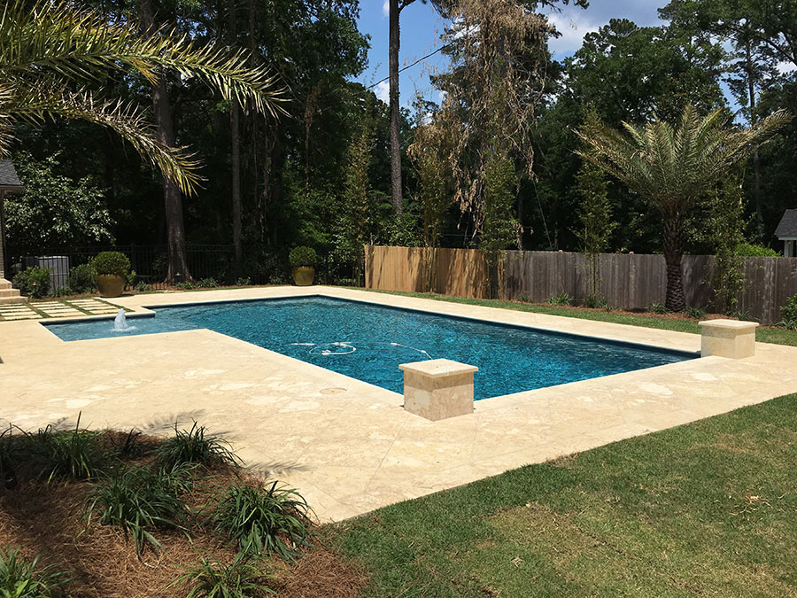 Mystic Pools pool renovation in Cairo featuring a rectangular pool with fountain jet, travertine paver decking, stone pedestal accents, and a wooden privacy fence with palm tree landscaping