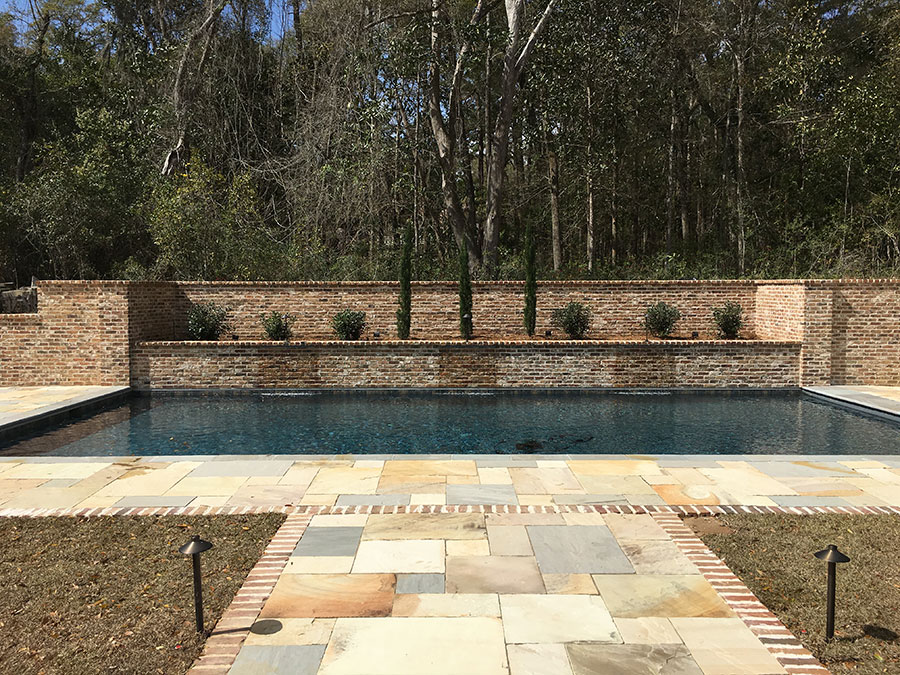 Mystic Pools custom saltwater gunite pool in Cairo with dark finish, raised brick retaining wall, flagstone paver patio with brick border, landscape path lighting, and Italian cypress and shrub plantings