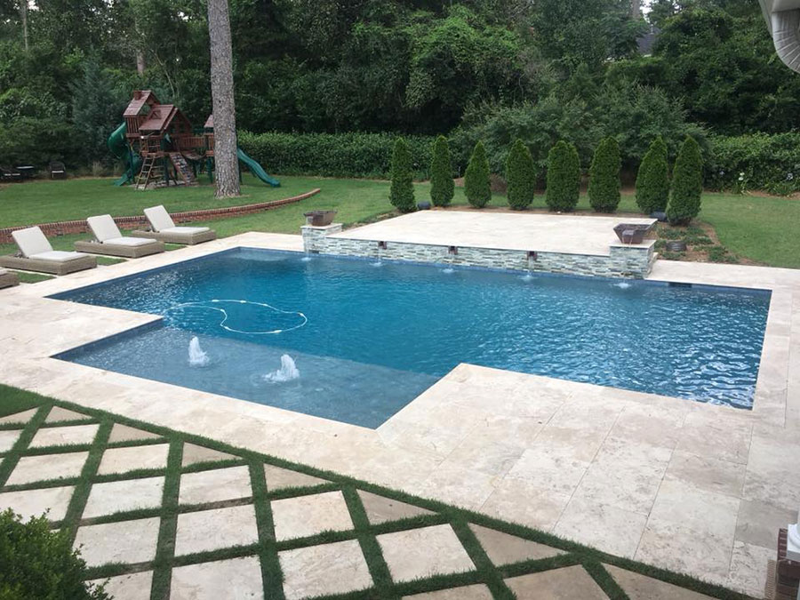 Mystic Pools custom rectangular gunite pool with dual fountain jets, raised stacked stone spillover wall with copper bowl accents, travertine paver decking, lounge chairs, and wooded backyard with children's play set
