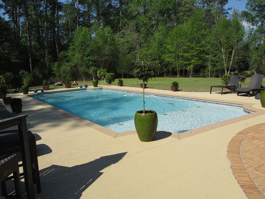 Mystic Pools custom pool design in Madison featuring a rectangular gunite pool with submerged tanning ledge, decorative potted tree planters, brick paver border, lounge chairs, and a concrete pool deck surrounded by wooded landscaping