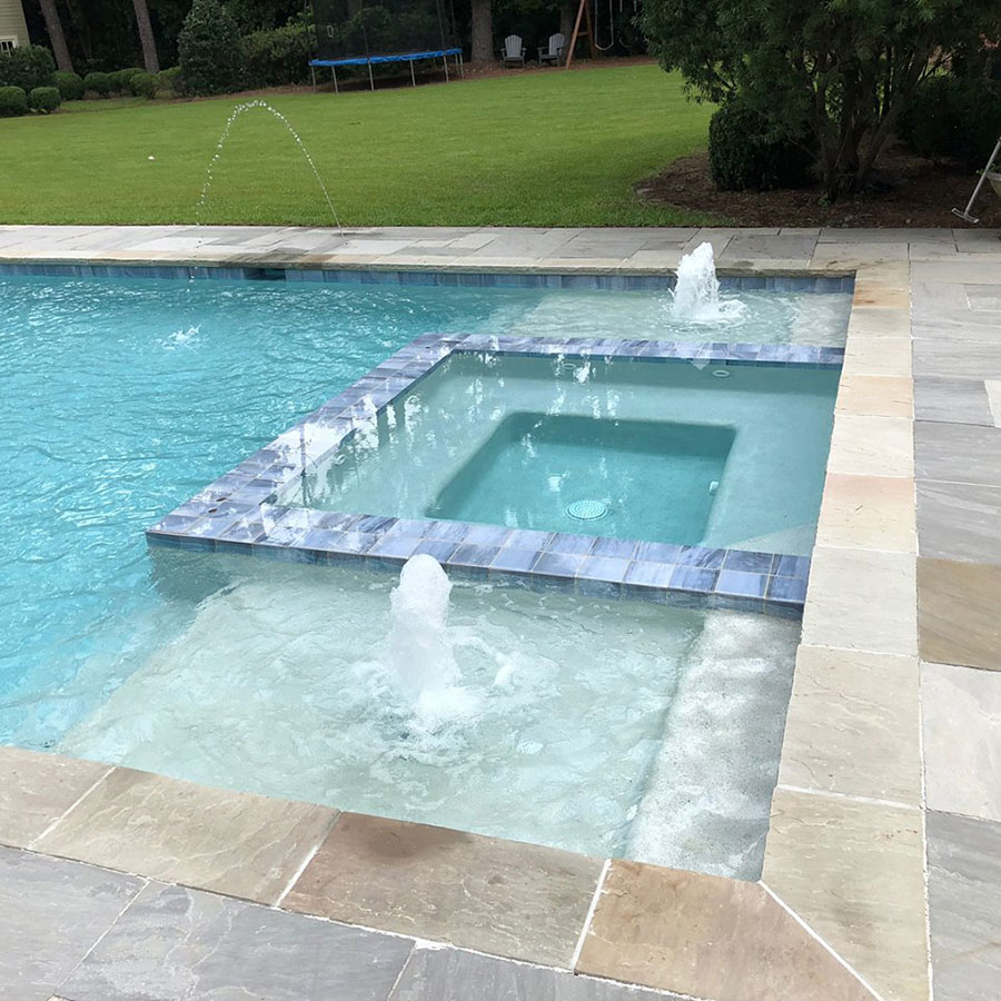 Mystic Pools custom gunite pool in Madison featuring a raised square spa with decorative tile, multiple fountain jets, zero-entry wading area, and natural stone paver decking