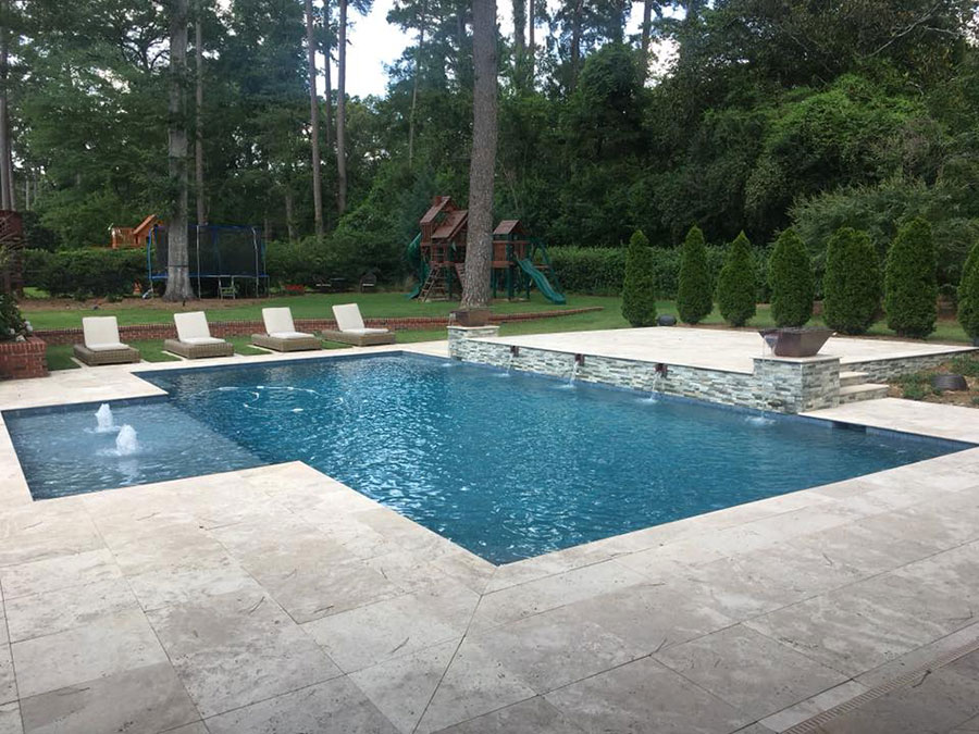 Mystic Pools custom rectangular gunite pool in Madison with fountain jet, raised stacked stone spillover wall, travertine paver decking, lounge chairs, and wooded backyard with children's play set