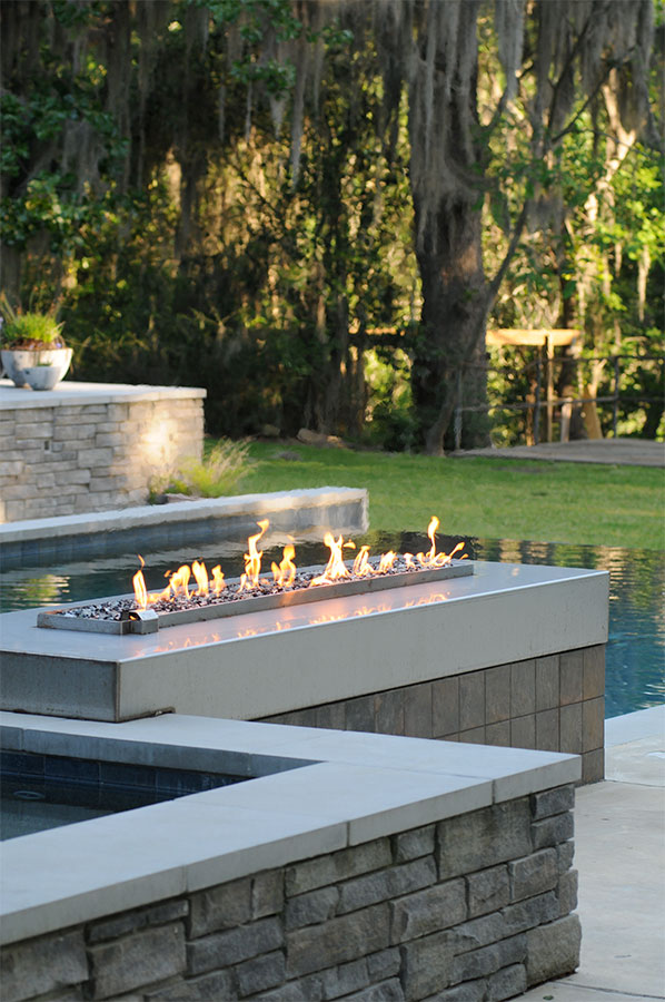 Close-up of Mystic Pools custom pool construction in Madison featuring a modern linear gas fire feature with glass rock media mounted on a stacked stone pool wall between the spa and main pool