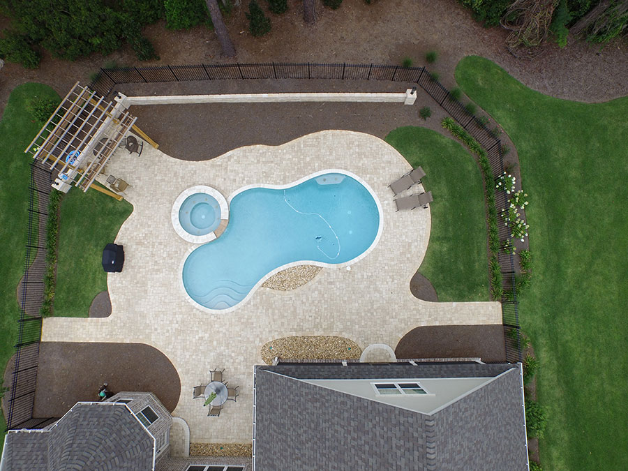 Aerial view of Mystic Pools pool renovation in Madison featuring a freeform figure-eight gunite pool with attached circular spa, travertine paver decking, wood pergola, dining area, and a fenced backyard surrounded by lawn and trees