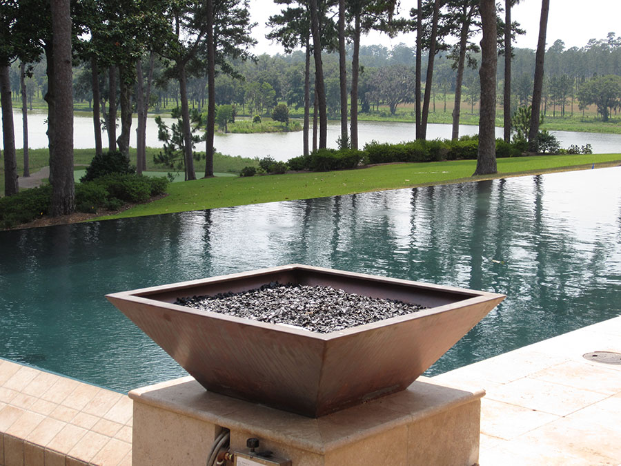 Mystic Pools remodeled infinity edge pool in Madison featuring a square copper fire bowl with glass rock media on travertine pedestal, with a serene lakefront and pine tree view reflected in the pool