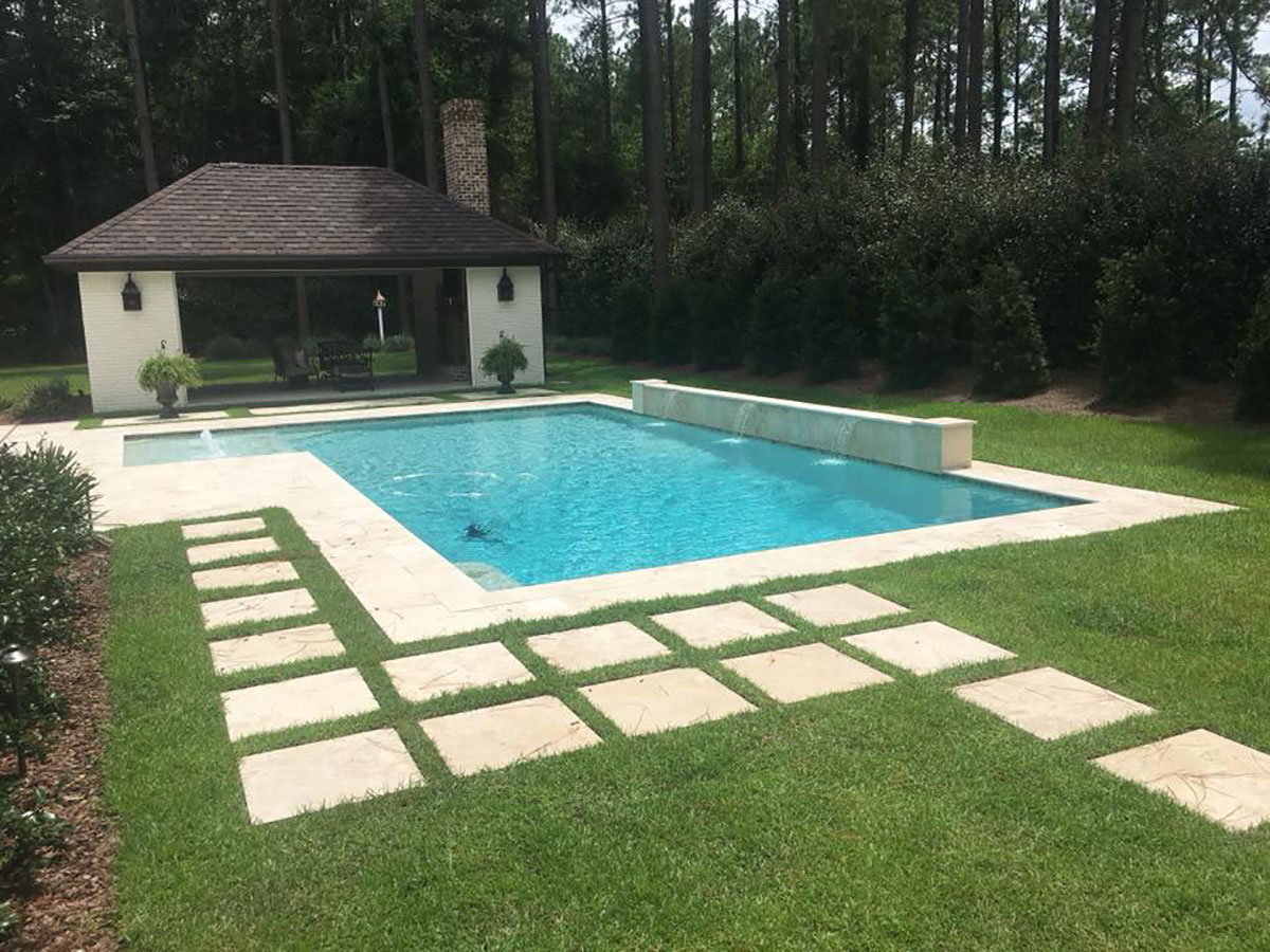 Mystic Pools custom saltwater gunite pool in Madison with raised spillover wall, travertine paver decking, grid stepping stone pathways, and a brick pool house with chimney surrounded by pine trees and hedges