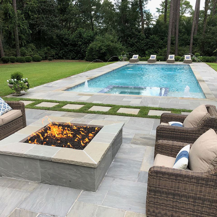 Mystic Pools custom rectangular gunite pool in Monticello with attached spa, fountain jets, arc water features, stone paver decking, built-in gas fire pit, and wicker outdoor seating surrounded by manicured landscaping