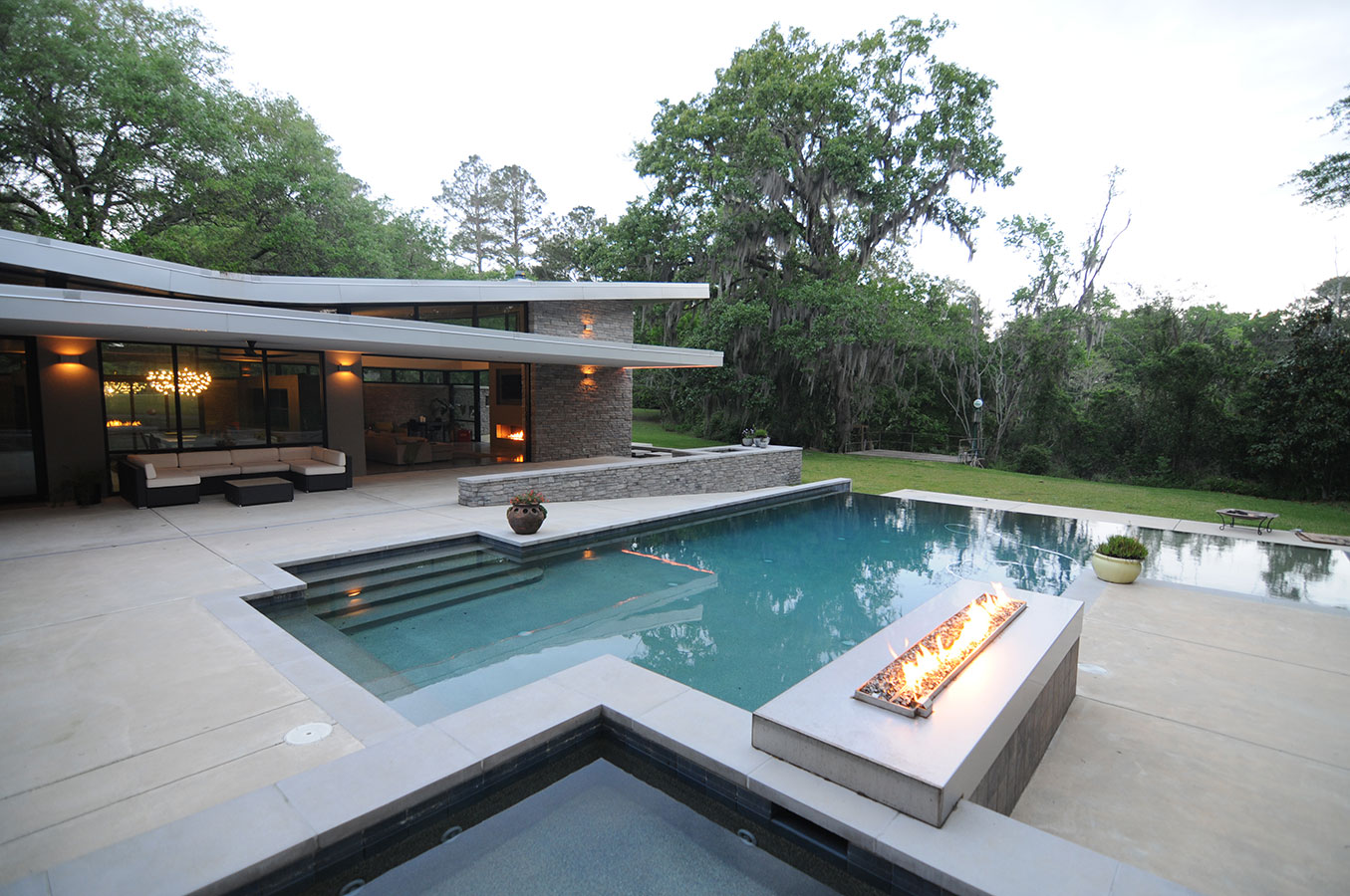 Mystic Pools custom gunite pool construction in Monticello featuring a modern geometric pool with dark tile finish, poolside linear gas fire feature, raised stacked stone spa wall, concrete deck, and a mid-century modern home with Spanish moss-draped oaks