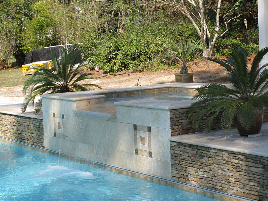 Mystic Pools pool renovation in Monticello featuring a raised travertine spa wall with decorative tile inlay accents, sheer waterfall spillover into the pool, and stacked stone base with sago palm landscaping
