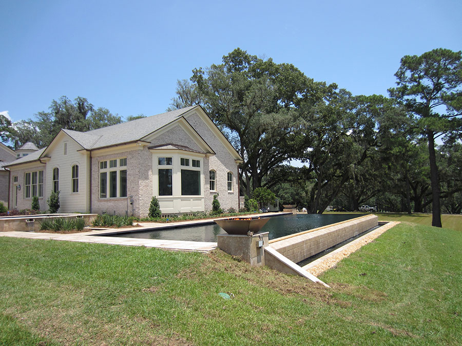 Mystic Pools remodeled rectangular lap pool in Monticello with dark finish, travertine coping, raised linear fire trough feature, and a white brick estate home surrounded by Spanish moss-draped live oaks
