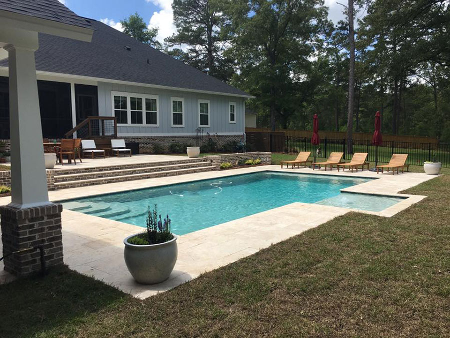 Mystic Pools residential gunite pool construction in Monticello featuring a rectangular pool with submerged tanning ledge, travertine paver decking, brick entry steps, teak lounge chairs with red umbrellas, and a board and batten home with covered porch