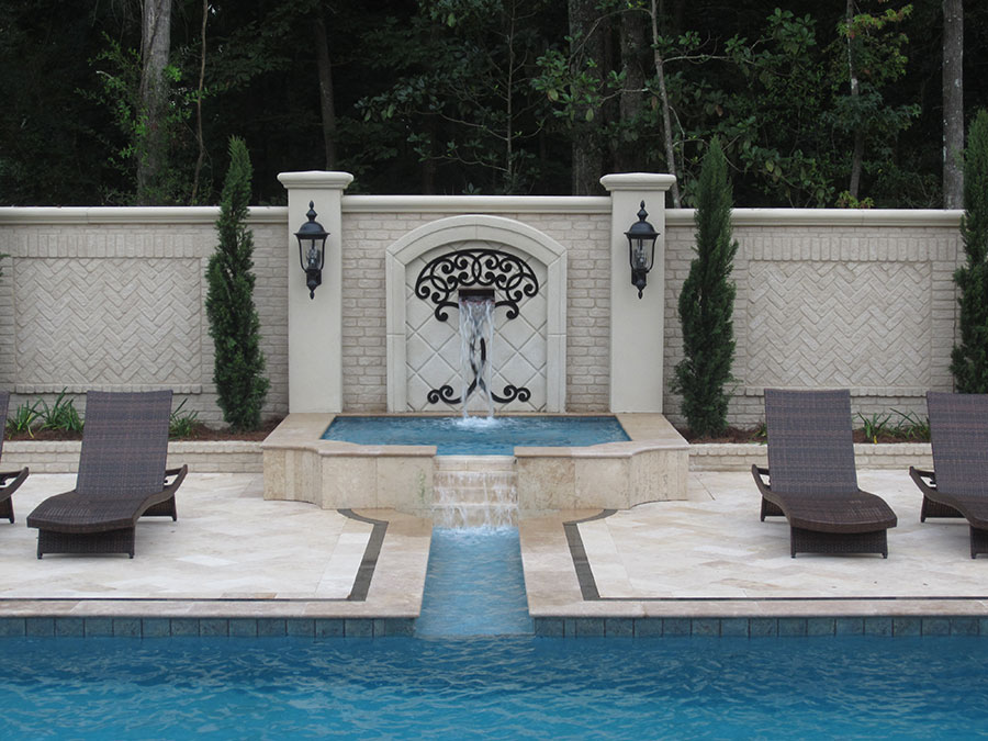 Mystic Pools custom gunite pool in Moultrie featuring a raised spa with ornate wrought iron wall fountain, cascading spillover into the main pool, herringbone brick accent wall, flanking columns with lanterns, and travertine paver decking with lounge chairs