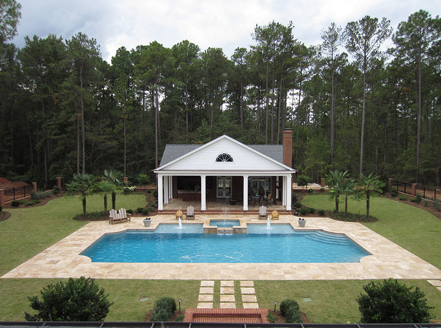 Aerial view of Mystic Pools custom rectangular gunite pool in Moultrie with attached spa, fountain jets, travertine paver decking, Adirondack seating, and a classic Southern pool house surrounded by tall pines and palm trees