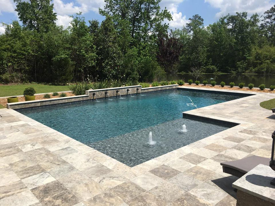 Mystic Pools custom gunite pool construction in Moultrie featuring a rectangular pool with raised wall scupper spillways, fountain jets, submerged tanning ledge, and travertine paver decking overlooking a tree-lined pond