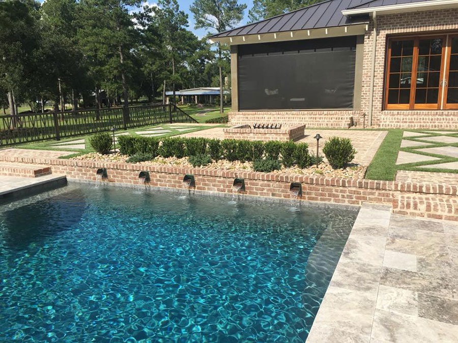 Mystic Pools pool renovation in Moultrie featuring a brick retaining wall with five scupper spillways, raised landscaped planter bed, travertine paver decking, and a brick home with retractable outdoor screen