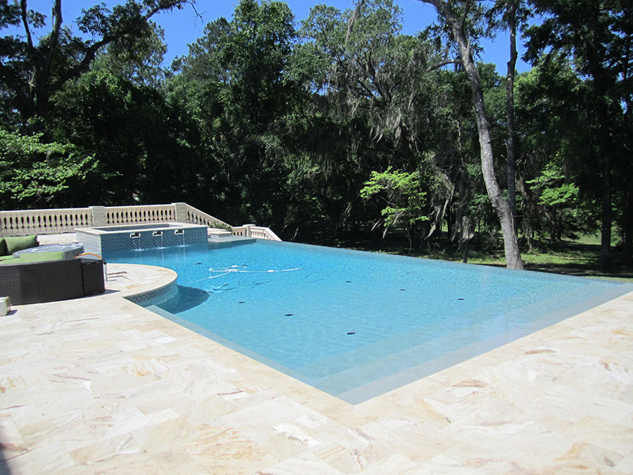 Mystic Pools custom infinity edge gunite pool in Quitman with attached spa, decorative balustrade, travertine paver decking, outdoor seating, and Spanish moss-draped trees in the background