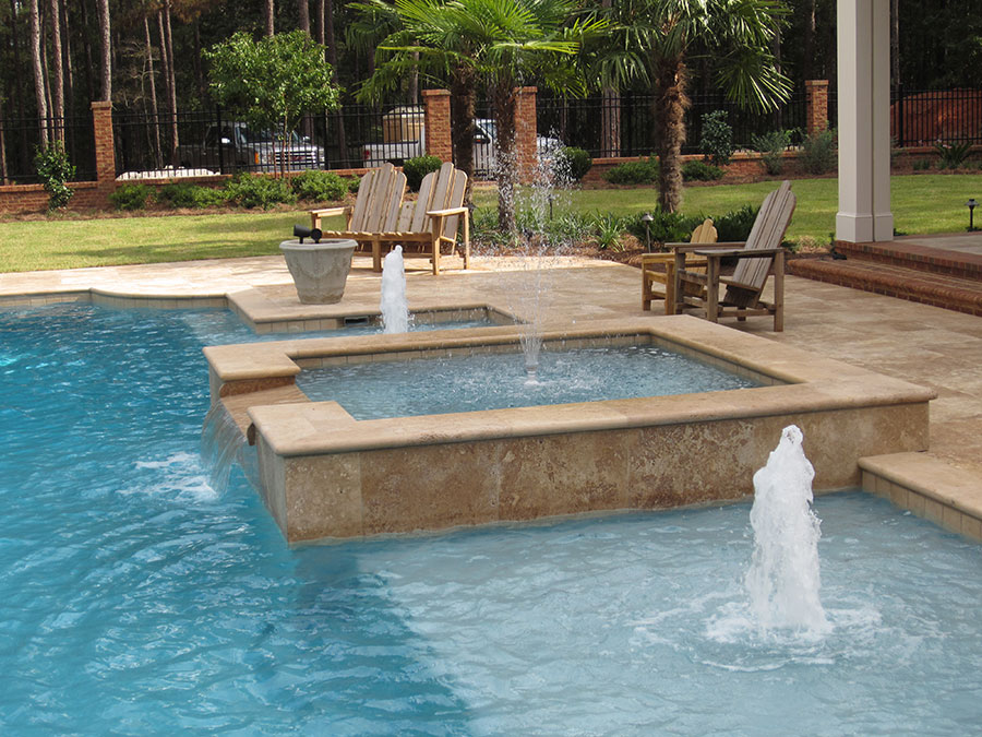 Mystic Pools custom gunite pool in Quitman featuring a raised square spa with travertine coping, cascading spillover, multiple fountain jets, Adirondack seating, and tropical palm tree landscaping with wrought iron fencing