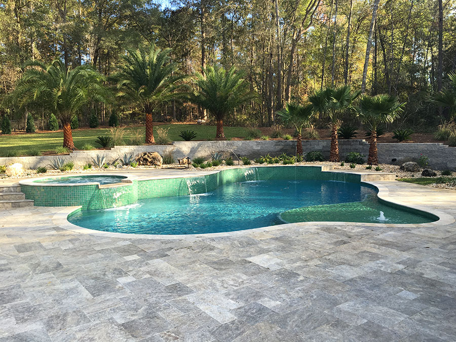 Mystic Pools custom freeform gunite pool construction in Quitman featuring green glass tile finish, circular raised spa with spillover waterfalls, travertine paver decking, and palm tree landscaping