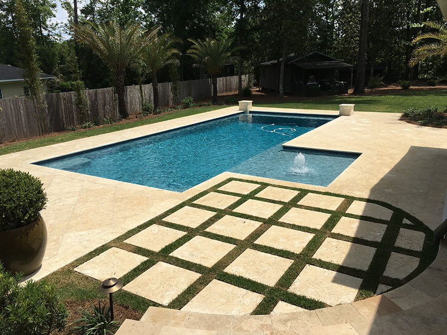 Mystic Pools pool renovation in Quitman featuring a rectangular pool with submerged tanning ledge, fountain jet, travertine paver decking, geometric grid paver and grass patio, and wood privacy fence with palm tree landscaping