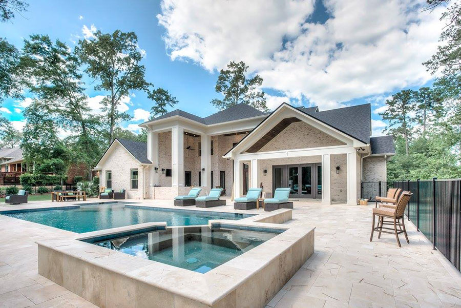 Mystic Pools remodeled rectangular gunite pool in Quitman with raised square spa, travertine paver decking, turquoise lounge chairs, covered outdoor patio, and a large stone and brick estate home surrounded by mature trees