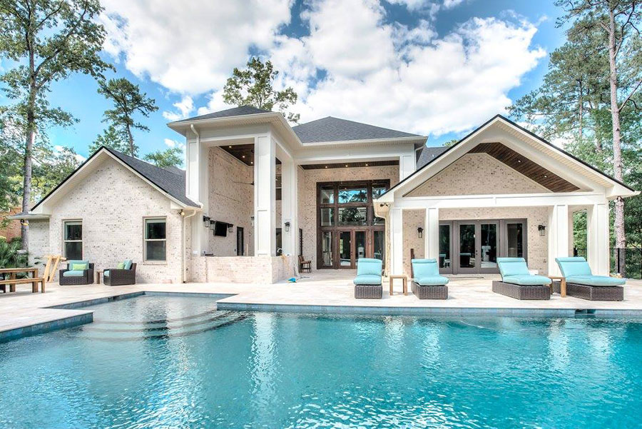 Mystic Pools custom saltwater gunite pool in Quitman with raised square spa, travertine paver decking, turquoise lounge chairs, covered outdoor patio, and a large stone and brick estate home surrounded by mature trees