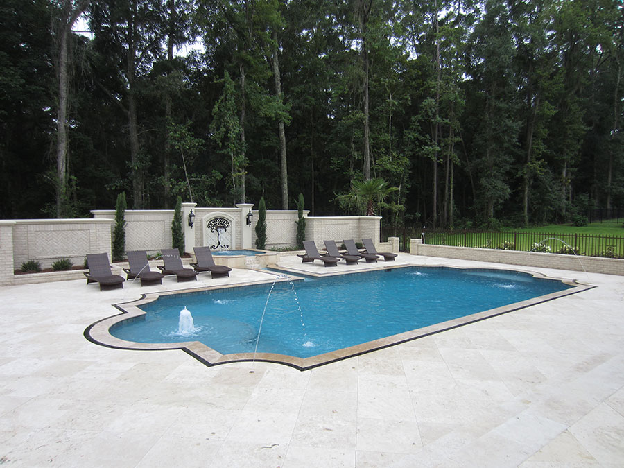 Mystic Pools custom freeform gunite pool in Saint George Island with fountain jet, travertine paver decking, attached spa, decorative brick accent wall with wrought iron lanterns, and lounge chairs surrounded by mature wooded landscaping