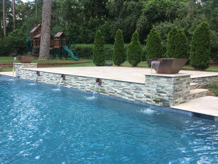 Mystic Pools custom gunite pool in Saint George Island featuring a stacked stone retaining wall with bronze scupper spillways, copper fire bowl accents, travertine coping, and evergreen arborvitae landscaping