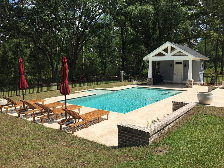 Mystic Pools custom gunite pool construction in Saint George Island featuring a rectangular pool with travertine paver decking, teak lounge chairs with red umbrellas, brick retaining wall, and a craftsman-style pool house surrounded by mature oak trees