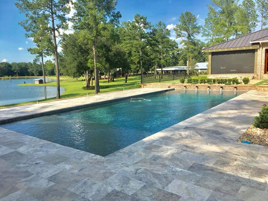 Mystic Pools pool renovation in Saint George Island featuring a rectangular infinity edge pool with dark finish, travertine paver decking, and a scenic lakefront view with a retractable outdoor screen and metal-roofed pool house