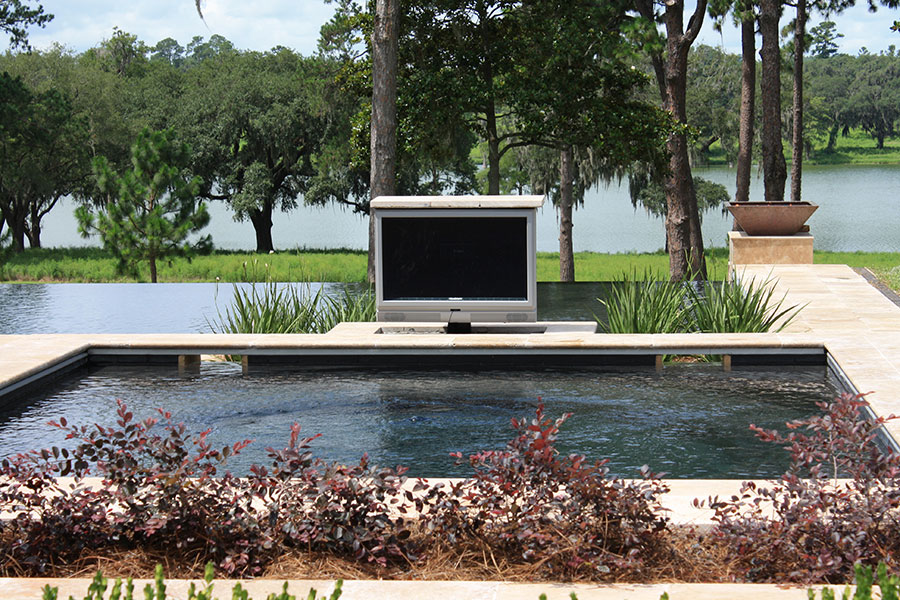 Mystic Pools remodeled infinity edge pool in Saint George Island with dark finish, poolside outdoor weatherproof TV, travertine coping, ornamental plantings, and a scenic lakefront view with Spanish moss-draped trees