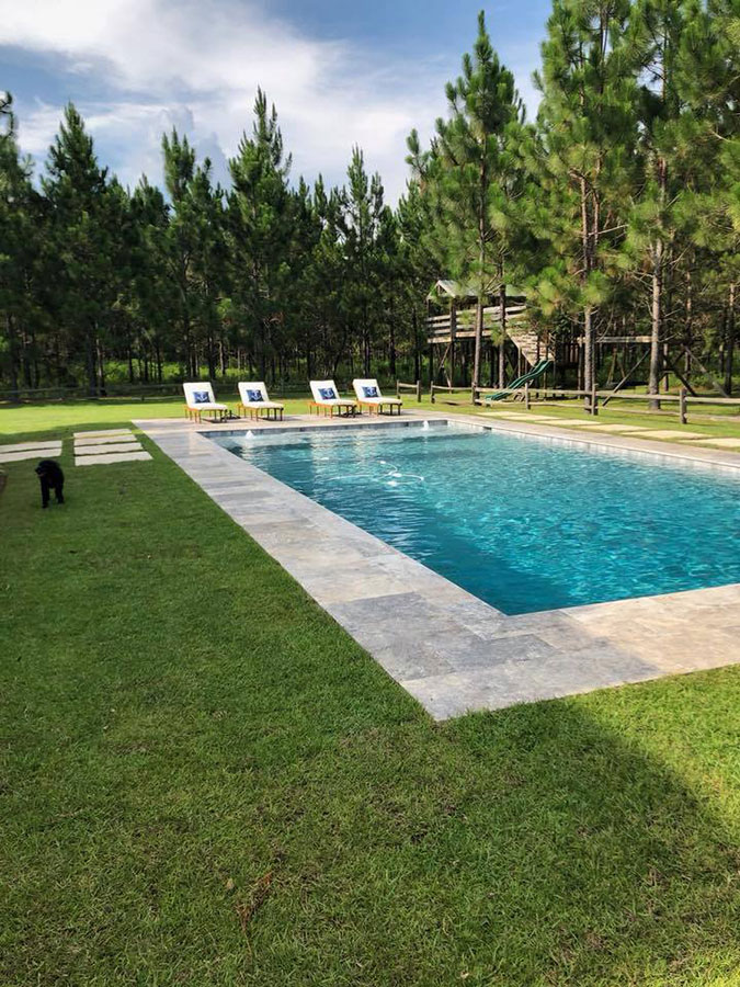 Mystic Pools custom saltwater gunite pool in Saint George Island with fountain jet, travertine paver decking, lounge chairs, stepping stone pathway, and an open lawn with a wooden play structure surrounded by tall pines