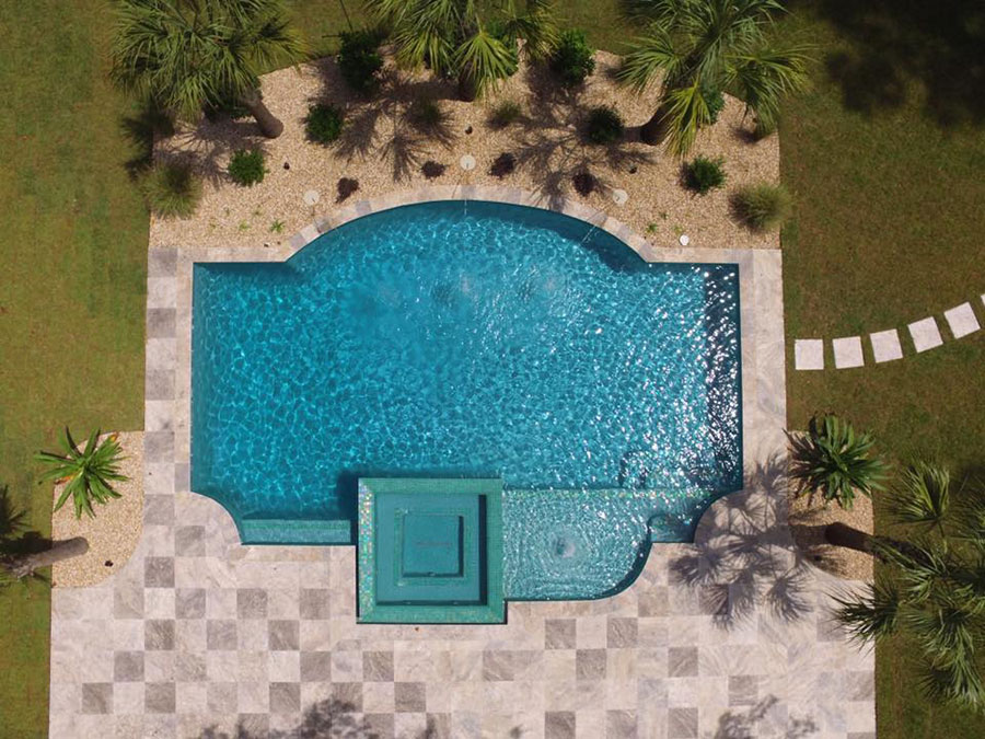 Aerial view of Mystic Pools custom gunite pool in Tallahassee with classic shaped pool, attached square spa with green glass tile, travertine paver decking, and tropical palm tree landscaping