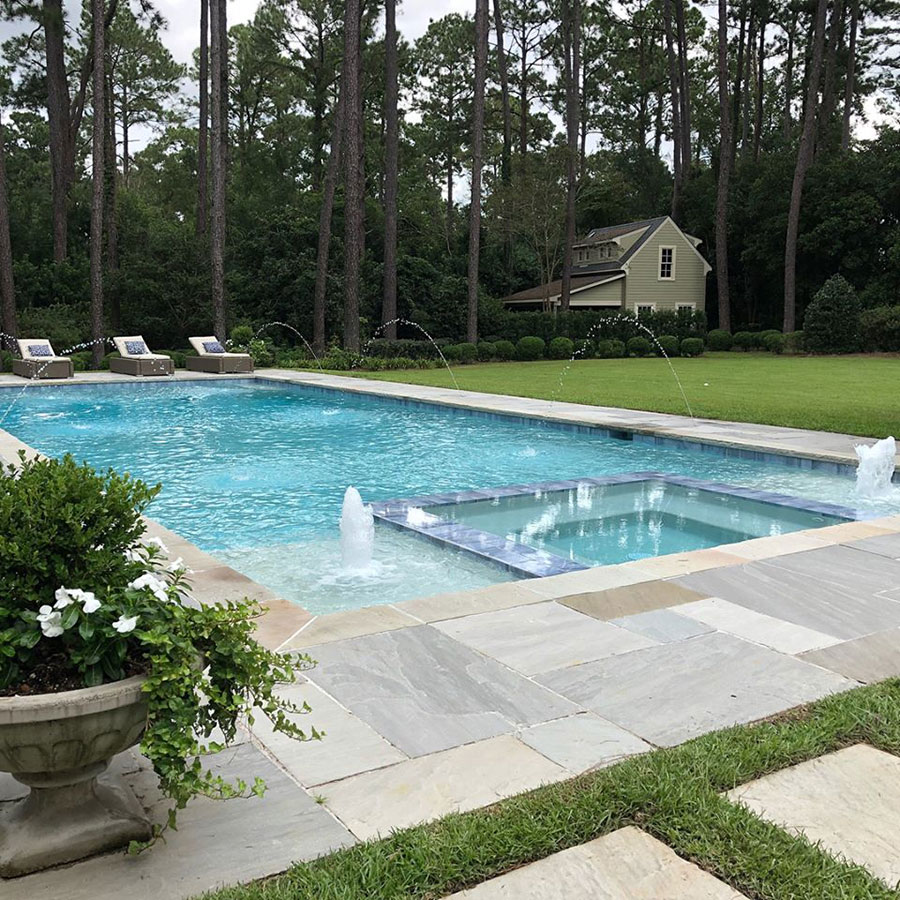 Mystic Pools custom rectangular gunite pool in Tallahassee with attached spa, center fountain jet, arc water features, natural stone paver decking, lounge chairs, and wooded backyard setting