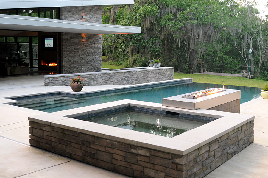 Mystic Pools custom gunite pool construction in Tallahassee featuring a modern rectangular pool with dark tile finish, raised stacked stone spa, concrete linear fire feature, and a contemporary stone and glass home with Spanish moss-draped trees
