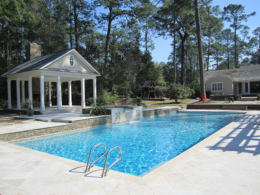 Mystic Pools pool renovation in Tallahassee featuring a rectangular pool with handrail, raised stacked stone wall with fountain feature, travertine paver decking, and an open-air pool pavilion with fireplace surrounded by tall pines