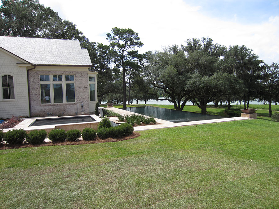Mystic Pools remodeled rectangular infinity edge pool in Tallahassee with dark finish, attached spa, travertine coping, manicured shrubs, and a scenic waterfront view framed by Spanish moss-draped live oak trees